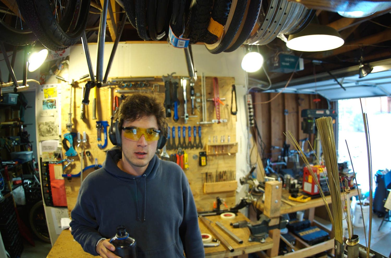 Spencer wearing PPE inside a workshop with tools on a plywood board, a workbench, and bike wheels hanging from the ceiling.